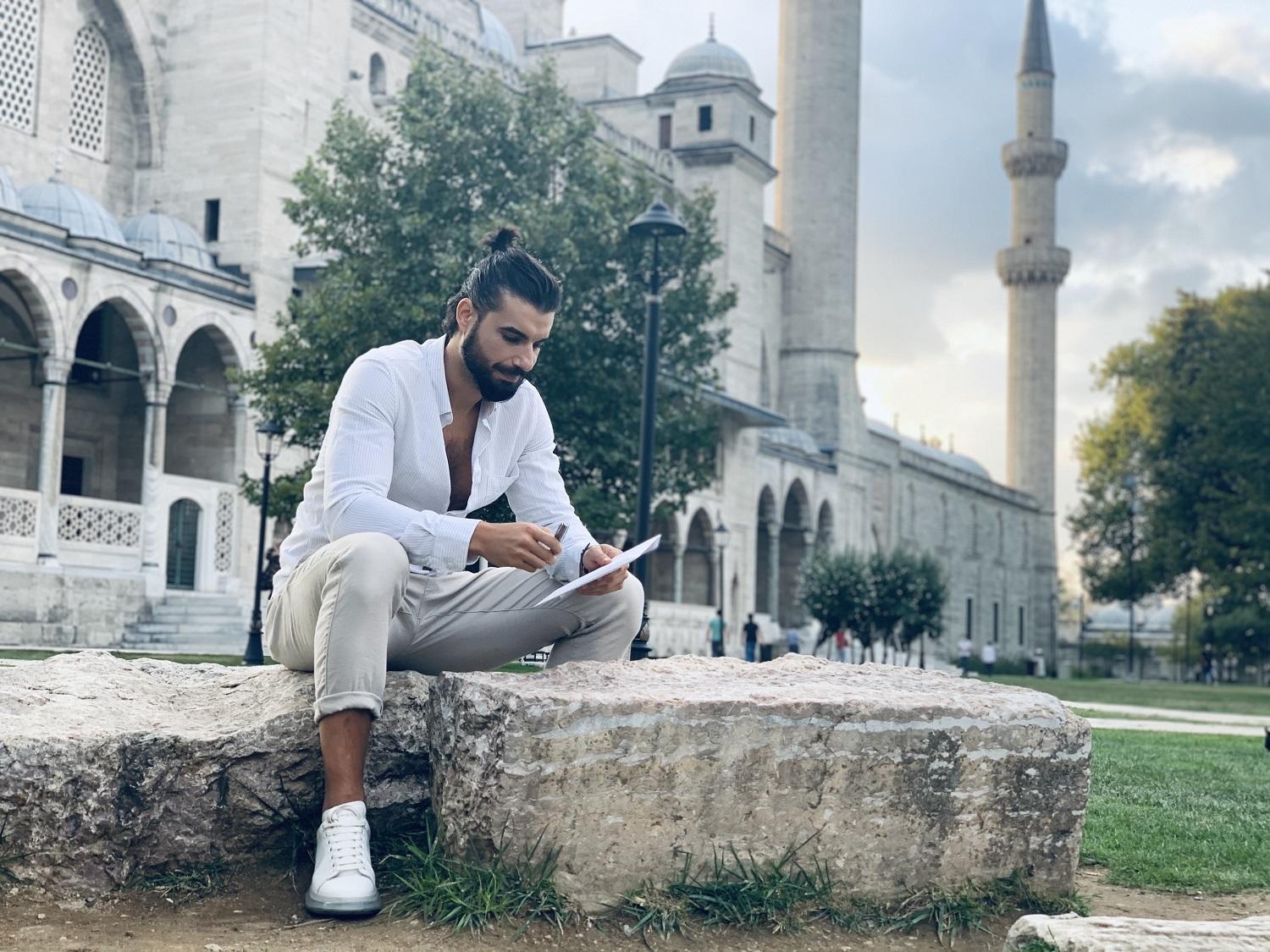 Andi Constantin într-o cămașă albă și pantaloni crem, stă pe piatră și scrie pe caiet