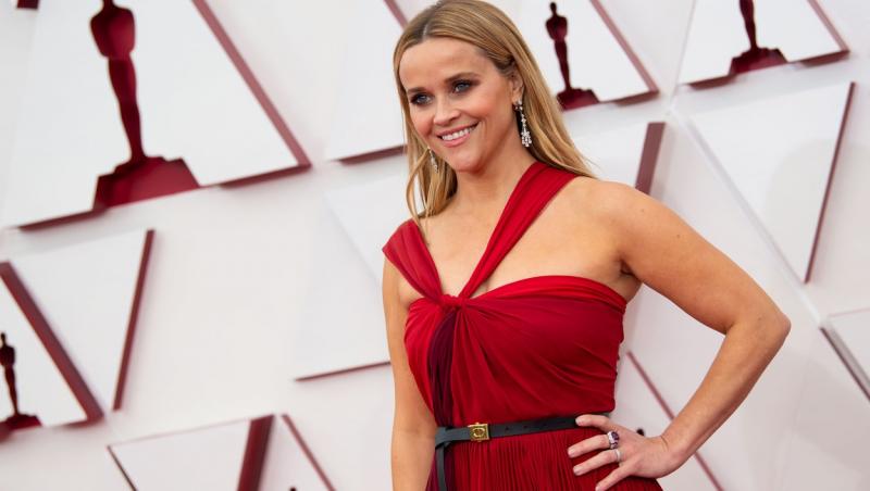 Reese Witherspoon, imbracata intr-o rochie rosie, tine mana in sold si este zambitoare