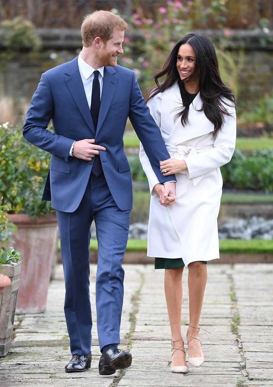 meghan markle intr-un palton alb si printul harry intr-un costum bleumarin tinandu-se de mana si uitandu-se unul la altul