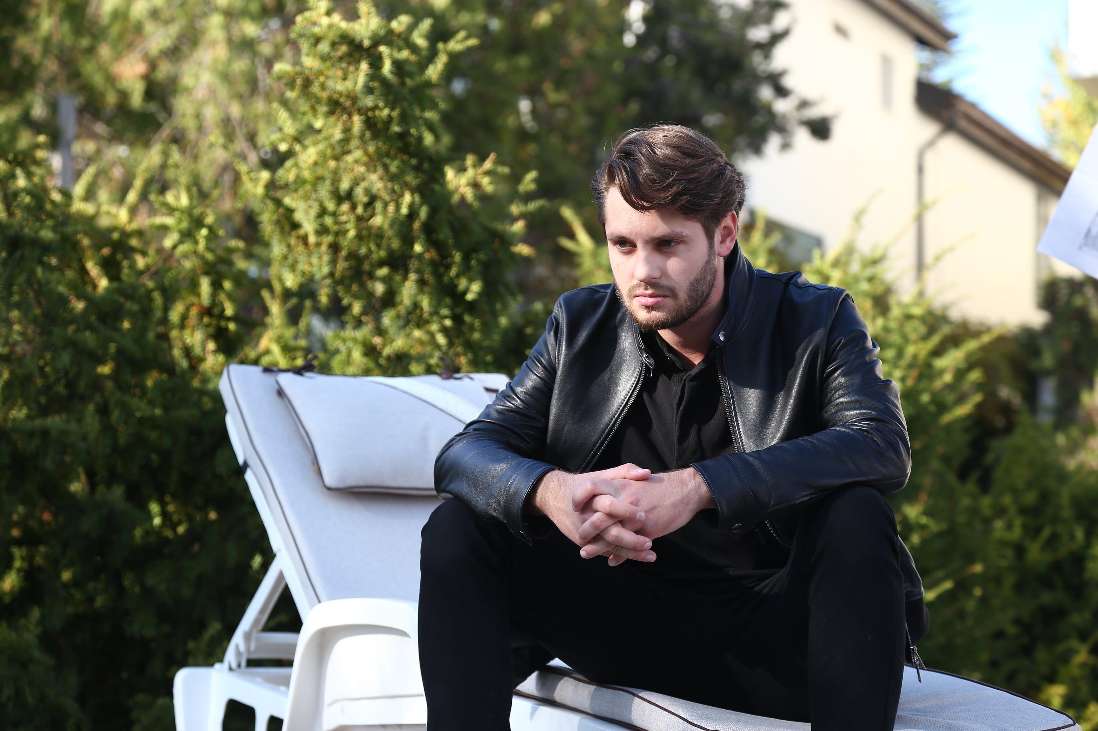 Luca, îngândurat, așezat pe un scaun de exterior, îmbrăcat în negru, cu o geacă de piele
