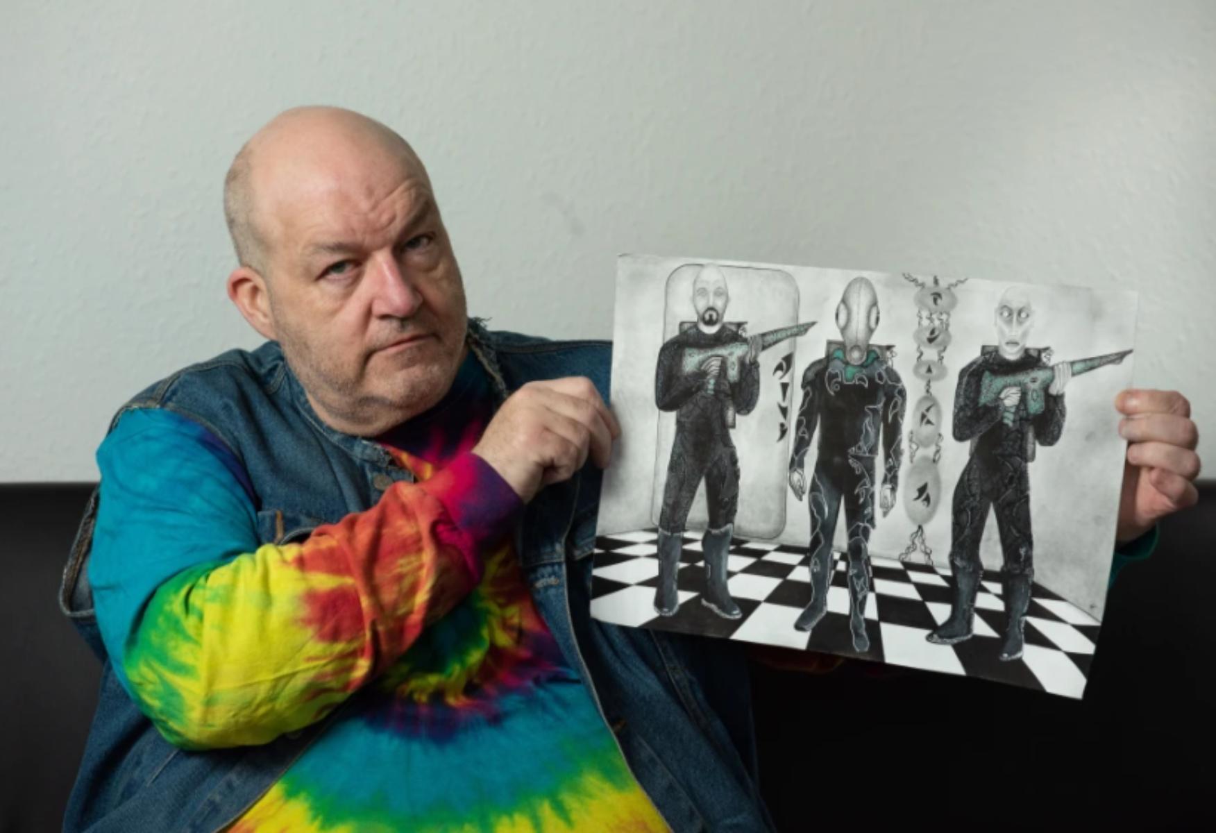 Bărbatul care susține că a fost răpit de extratereștri de 60 de ori, îmbrăcat cu un janorac colorat și arată un desen în creion cu trei entități extraterestre