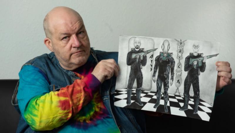 Bărbatul care susține că a fost răpit de extratereștri de 60 de ori, îmbrăcat cu un janorac colorat și arată un desen în creion cu trei entități extraterestre