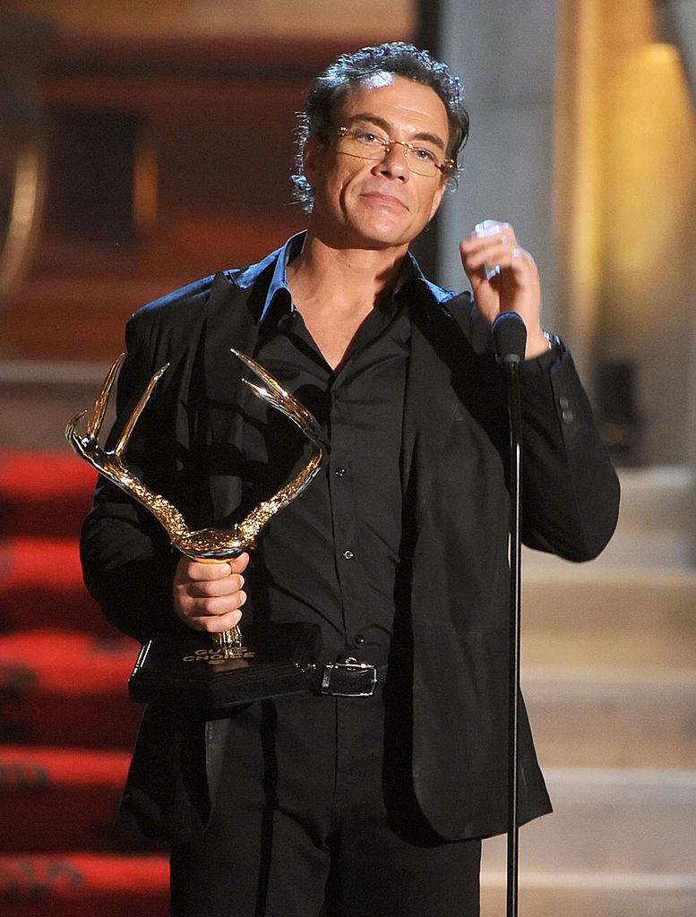 Jean-Claude-Van-Damme-cămașă-neagră-sacou-negru-ține-premiu-în-mână