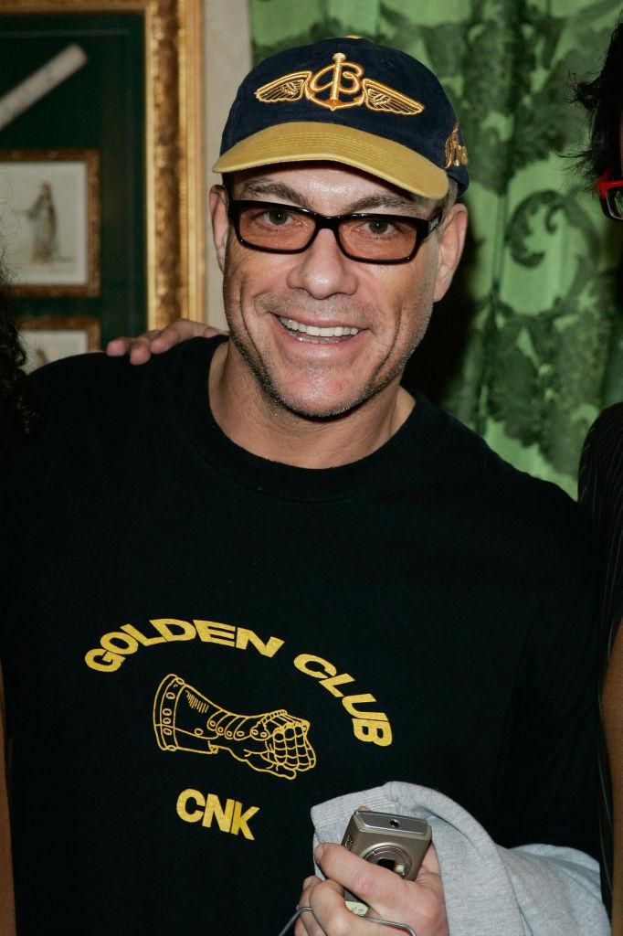 Jean-Claude-Van-Damme-bluză-neagră-șapcă-neagră-ochelari-zâmbește