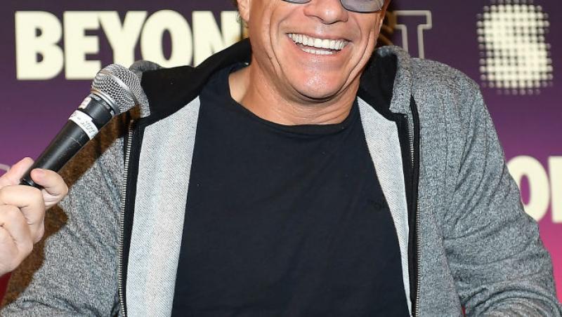 Jean-Claude-Van-Damme-hanorac-gri-zâmbește-șapcă-ochelari-stă-pe-scaun