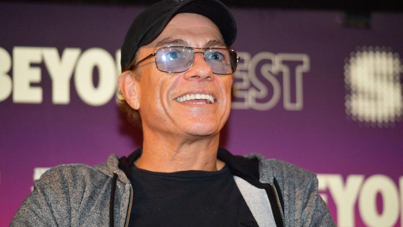 Jean-Claude-Van-Damme-hanorac-gri-zâmbește-șapcă-ochelari