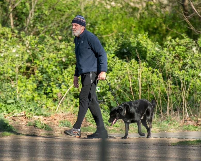 Hugh Laurie, la plimbare pe malul lacului, imbracat într-o bluză albastră, o pereche de pantaloni negri și căciulă albastră în dungi, supărat, împreună cu cățelul lui negru