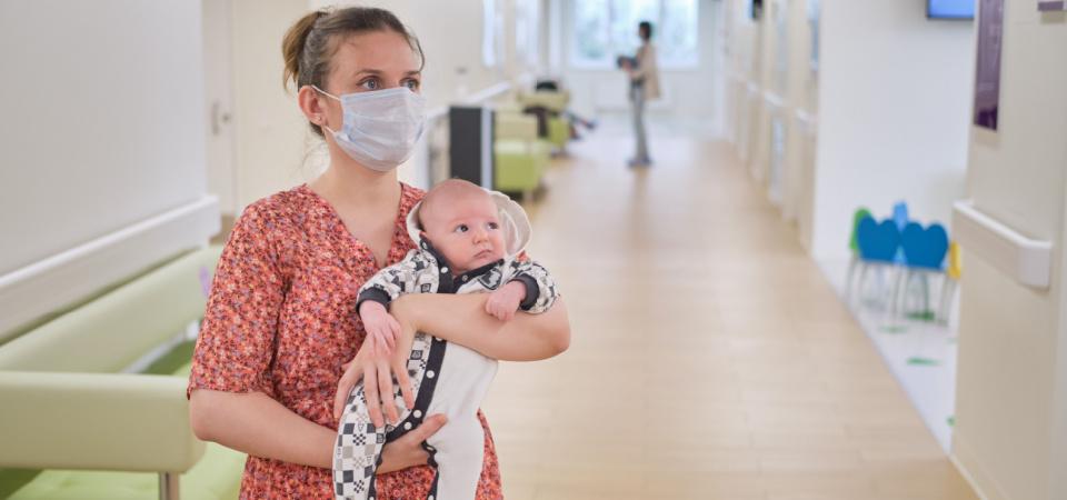 Imagine cu o mamă care își ține bebelușul în brațe în timp ce așteaptă să intre în cabinetul medicului.