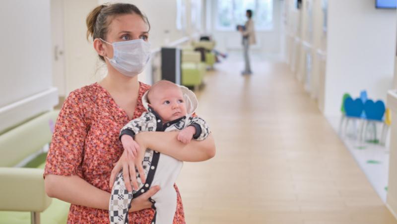 Imagine cu o mamă care își ține bebelușul în brațe în timp ce așteaptă să intre în cabinetul medicului.