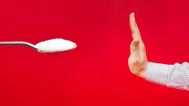 Imagine cu o lingură de zahăr și cu o mână a unei persoane care exprimă refuzul. Imaginea are fundal roșu.
