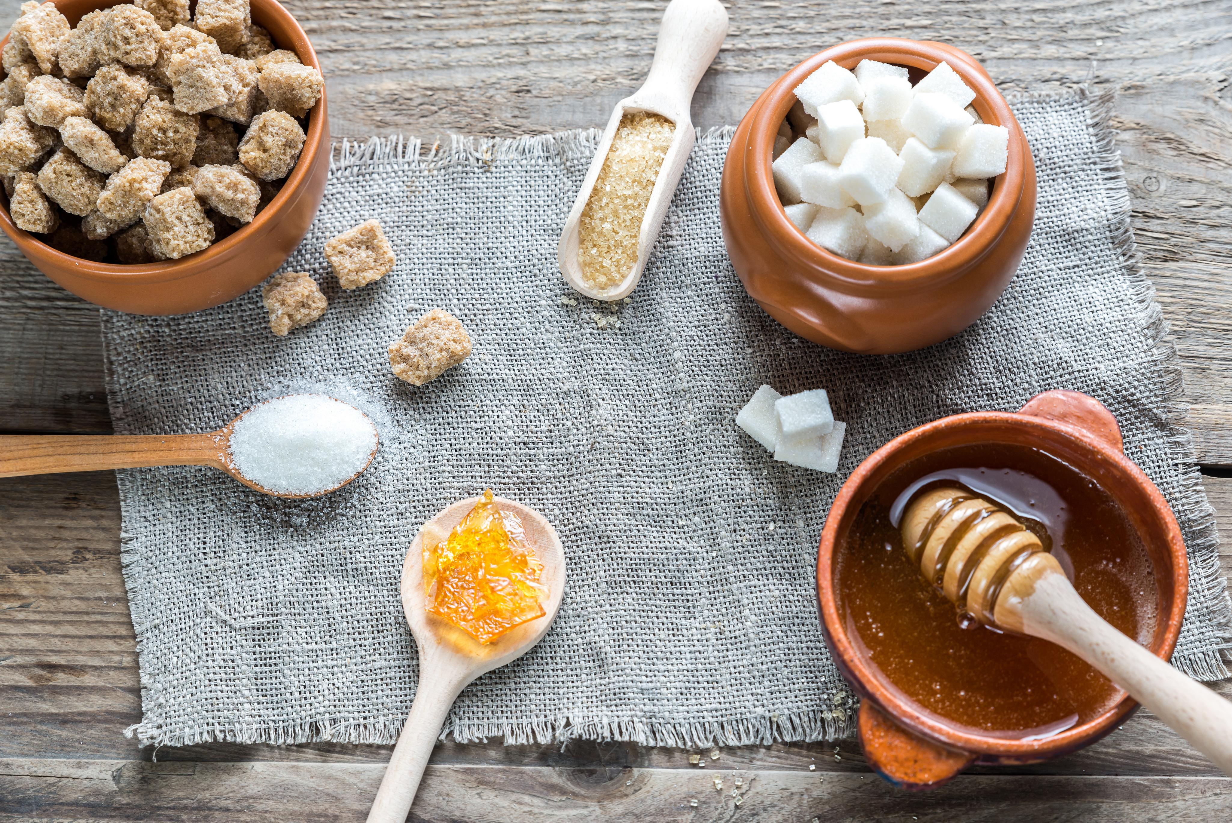Imagine cu o masă unde sunt amplase ingredientre precum zahăr, zahăr brun în diferite forme și miere.