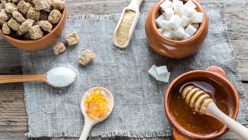 Imagine cu o masă unde sunt amplase ingredientre precum zahăr, zahăr brun în diferite forme și miere.
