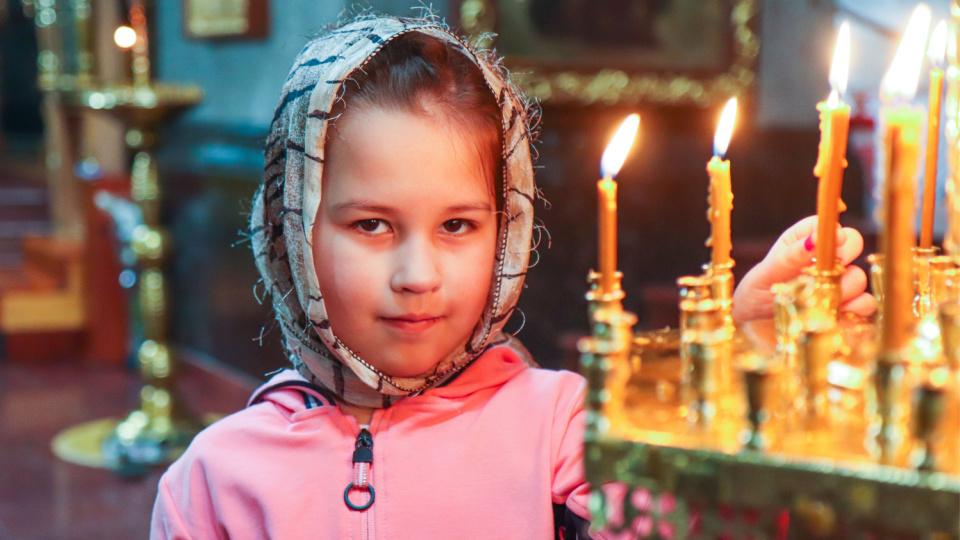 Imagine cu o fetiță care este la biserică și ține o lumânare aprinsă.