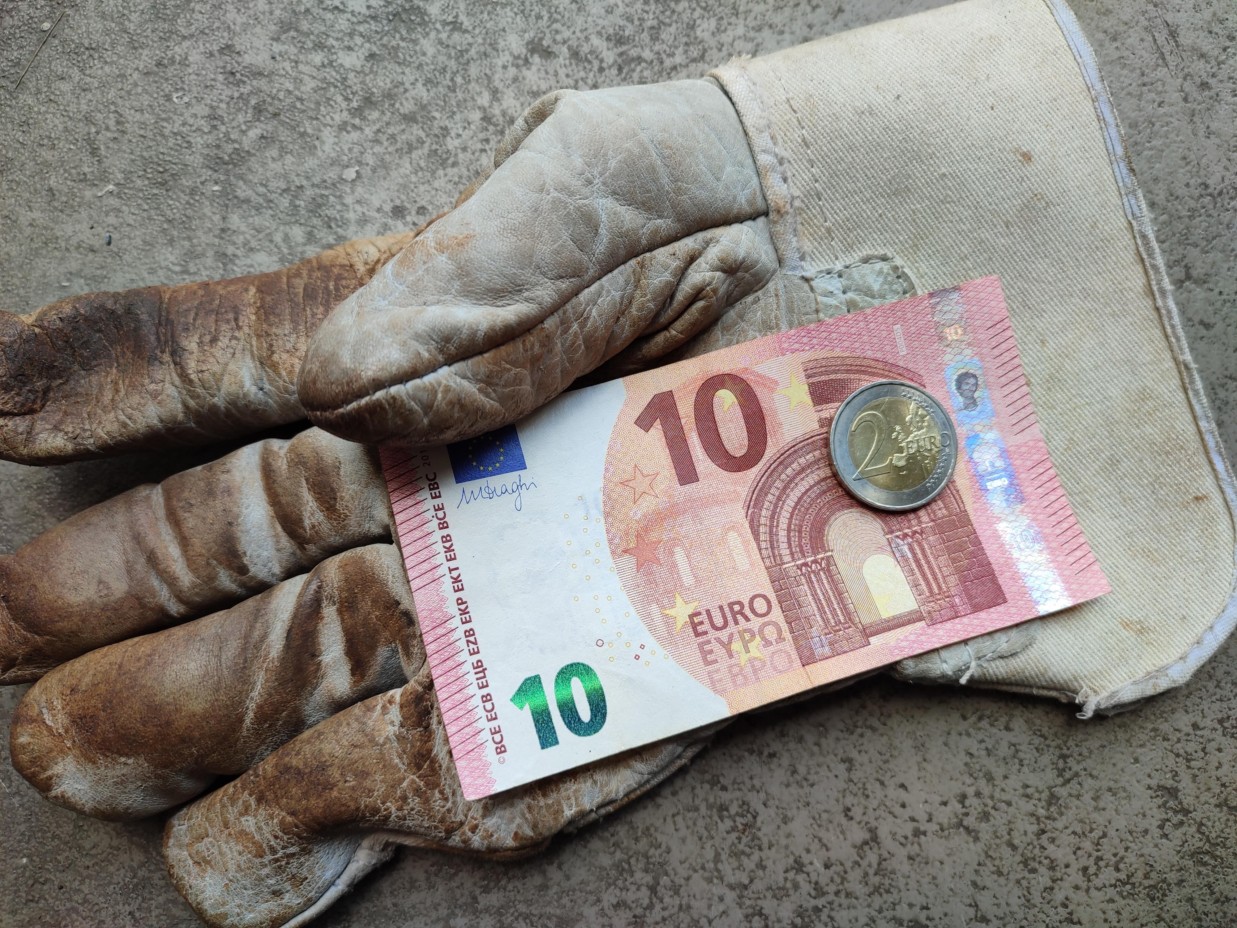 Imagine cu o mânușă murdară a unui om care a muncit și a primit o bancnotă de 10 euro și o monedă de 2 euro.