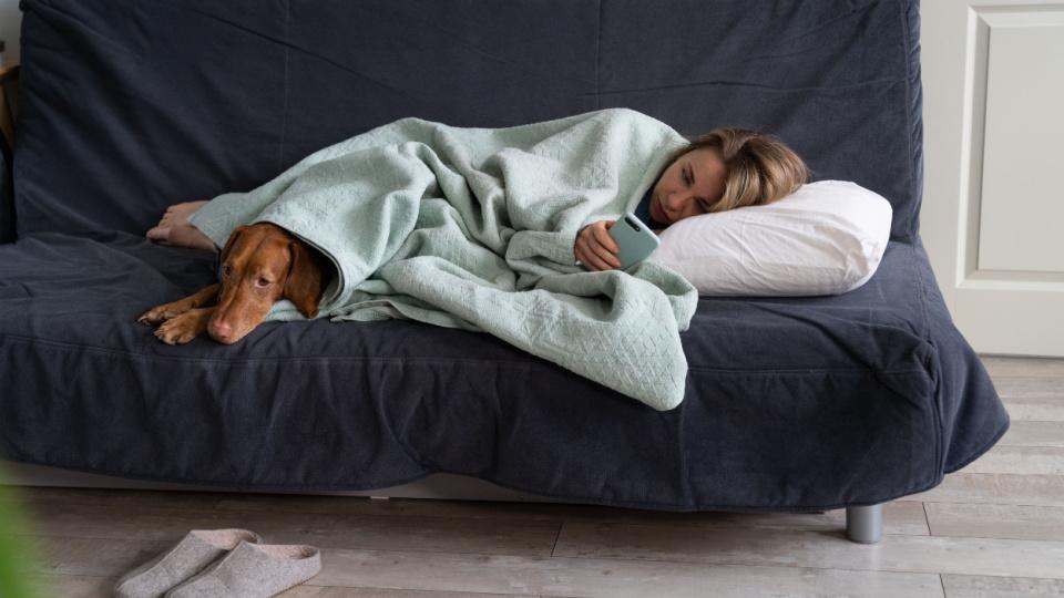 Tânără obosită și afectată de trecerea la ora de iarnă care stă întinsă pe canapea învelită alături de un cățel