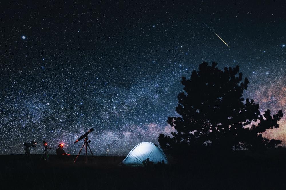 bărbat cu telescop stă pe scaun lângă cort și provește cerul în timpul unei ploi de meteori