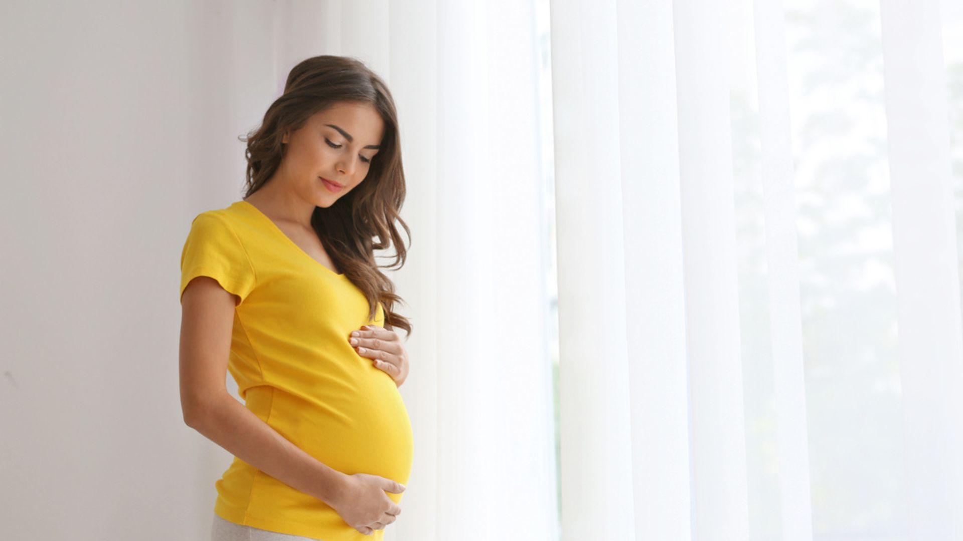 o mamă, îmbrăcată cu un tricou galben muștar, care își mângâie burta de gravidă
