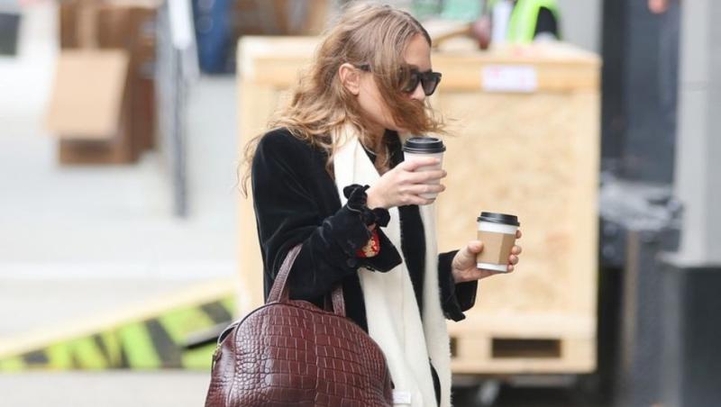 Ashley Olsen pe stradă cu un palton negru, o rochie roșie, o eșarfă albă. Are în mână o geantă din piele și o cafea