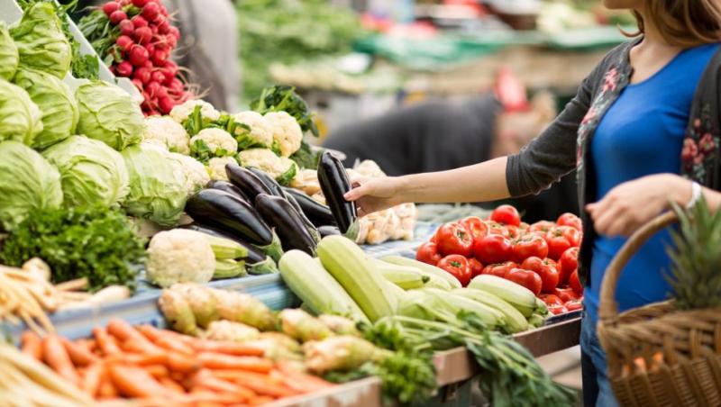 femeie care alege legume la piață: varză, vinete, morcovi, dovlecei și are în mână un coș