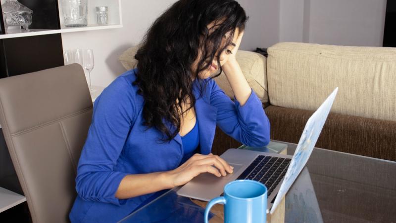 femeie în albastru care își ține capul într-o mână și se uită tristă la laptop