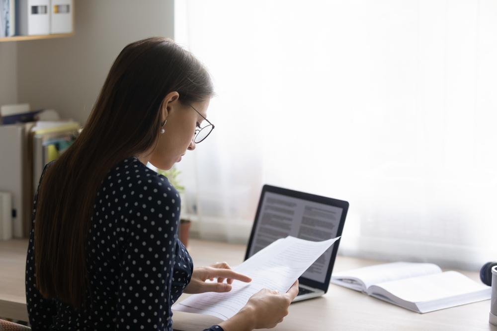 tânără cu ochelari și bluză închisă la culoare stând la birou cu o foaie în mână șângă o carte și un laptop