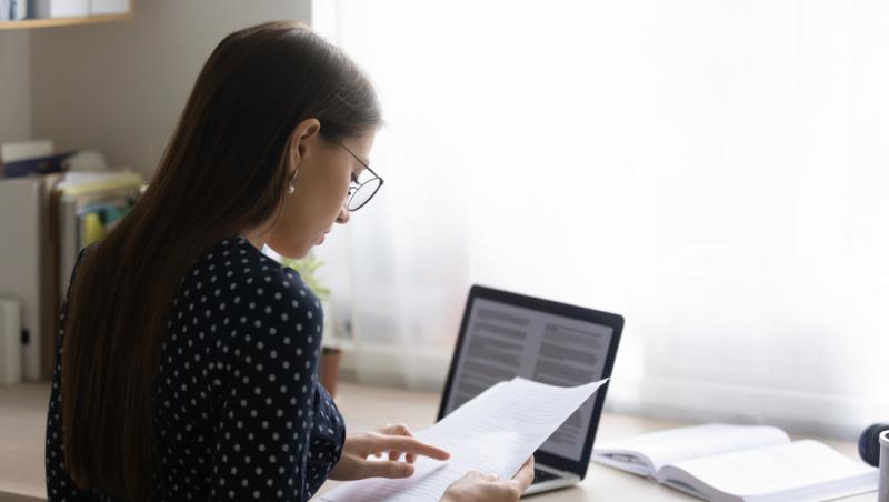 tânără cu ochelari și bluză închisă la culoare stând la birou cu o foaie în mână șângă o carte și un laptop