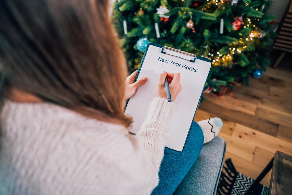 tânără cu părul lung și pulover alb care stă pe canapea cu un clipboard și un pix pentru a scrie rezoluțiile pentru noul an