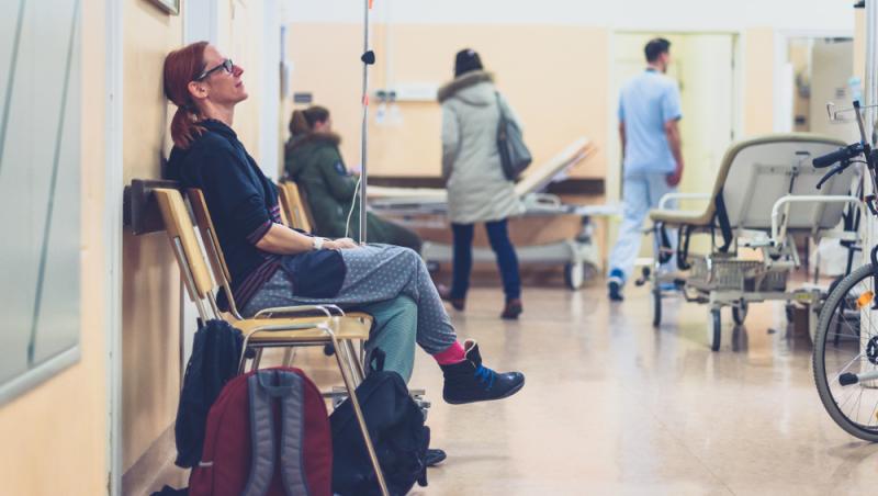 femeie în prim-plan care stă pe scaun de spital cu rucsac la picioare și așteaptă să intre la medic, iar în planul secund alți oameni și asistente