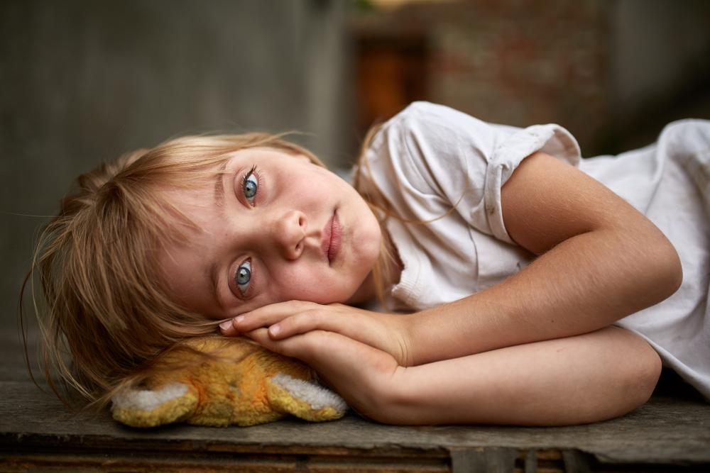 fetiță blondă cu ochi albaștri care stă cu mânuțele sub cap pe o jucărie de pluș murdară