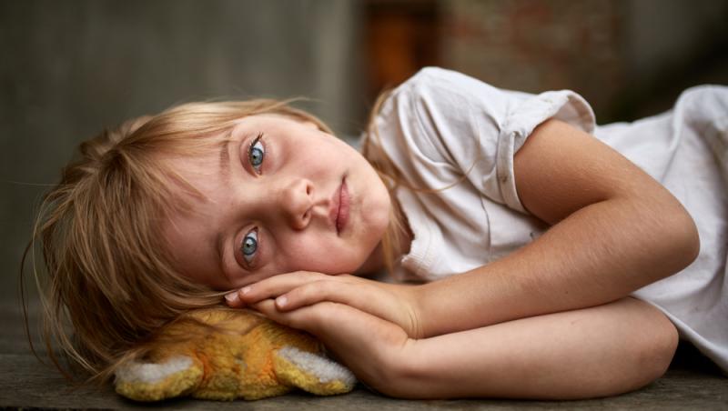 fetiță blondă cu ochi albaștri care stă cu mânuțele sub cap pe o jucărie de pluș murdară