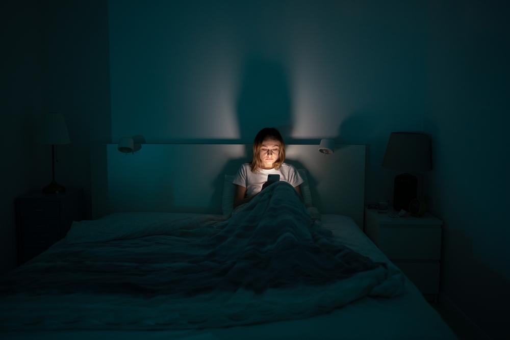 tânără care se uită pe telefon în timp ce stă pe întuneric în pat
