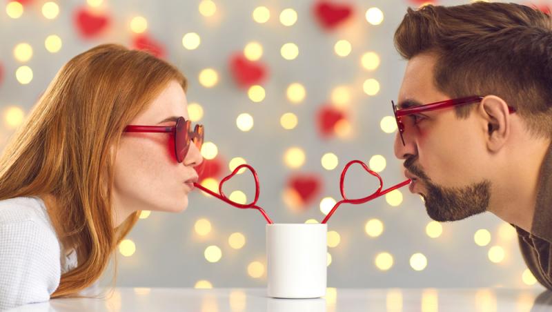 femeie și bărbat amândoi cu ochelari roșii, bând cu baie diferite dintr-o cană albă, iar pe fundal sunt luminițe și inimioare