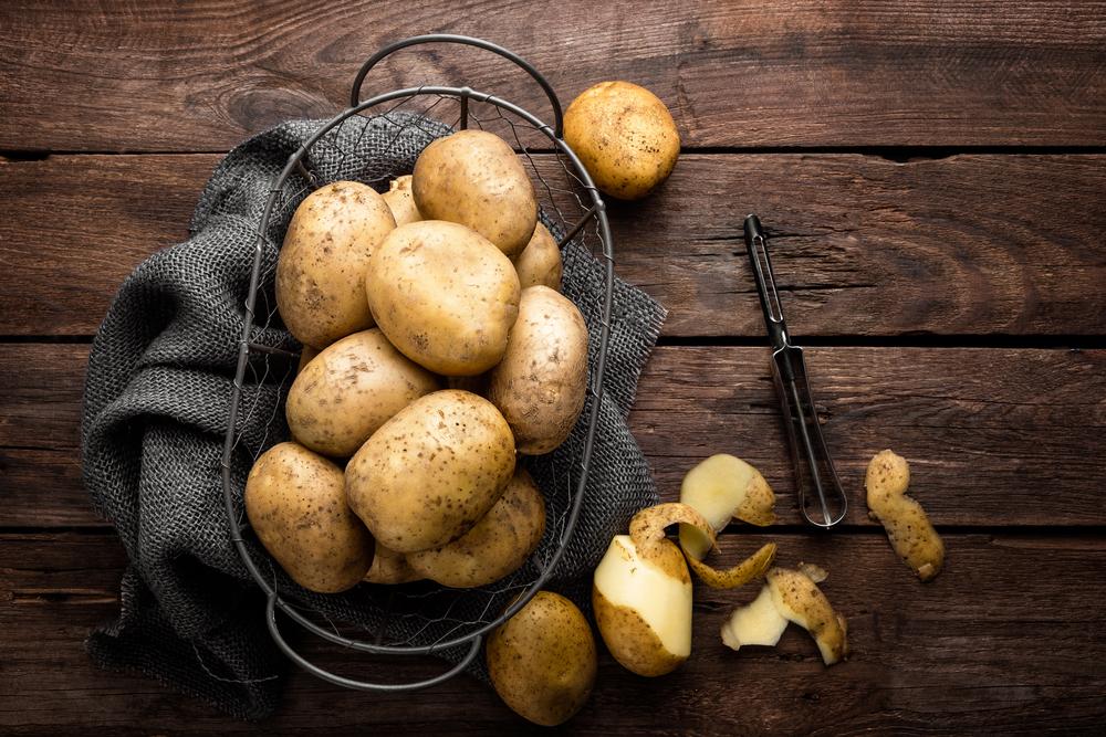 imagine cu niste cartofi varsati dintr-un sac pe o masa de lemn si niste coji de cartofi langa