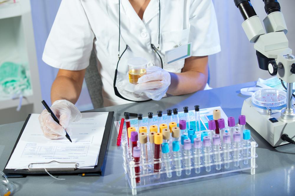 medic făcând analize medicale, notând într-o fișă și ținând în mână o mostră de urină, ar pe birou având mai multe eprubete cu sânge