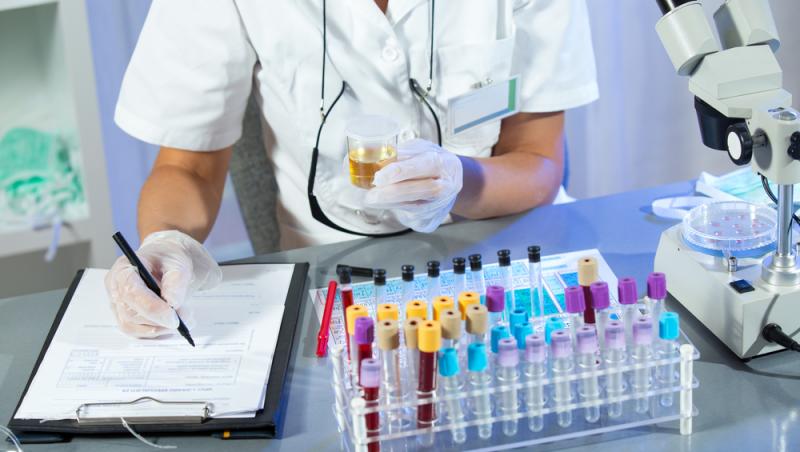 medic făcând analize medicale, notând într-o fișă și ținând în mână o mostră de urină, ar pe birou având mai multe eprubete cu sânge