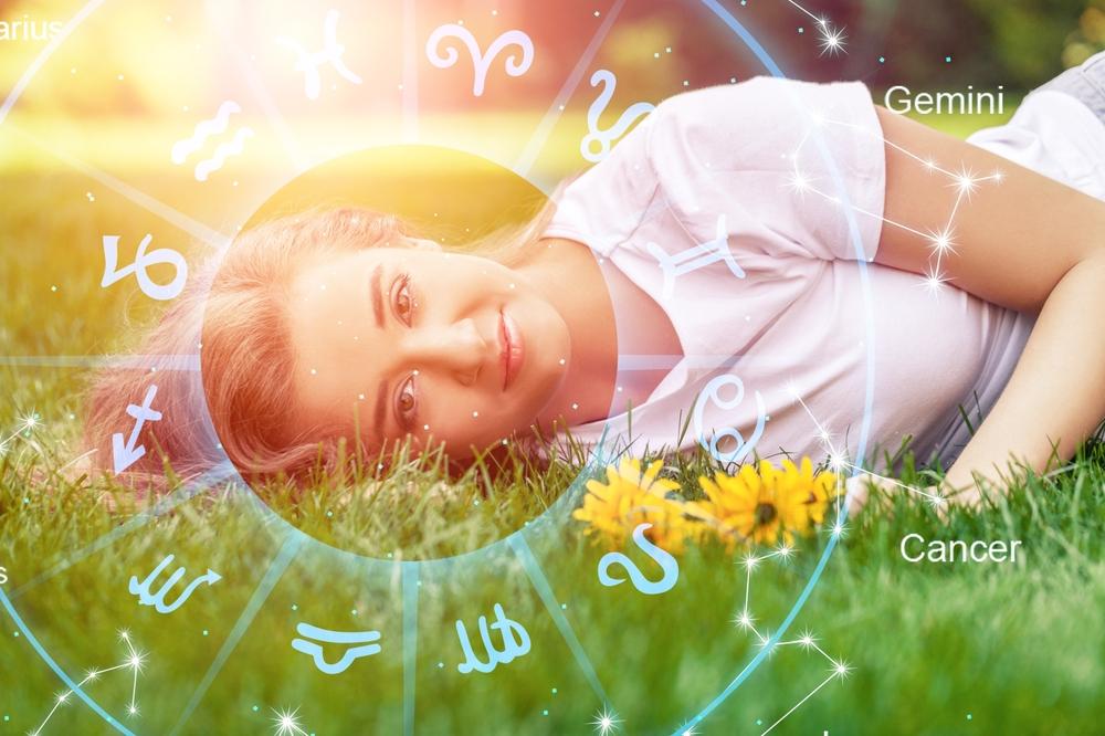 tânără așezată în iarbă lângă niște flori galbene și semnele zodiacale dispuse în cerc în jurul capului ei