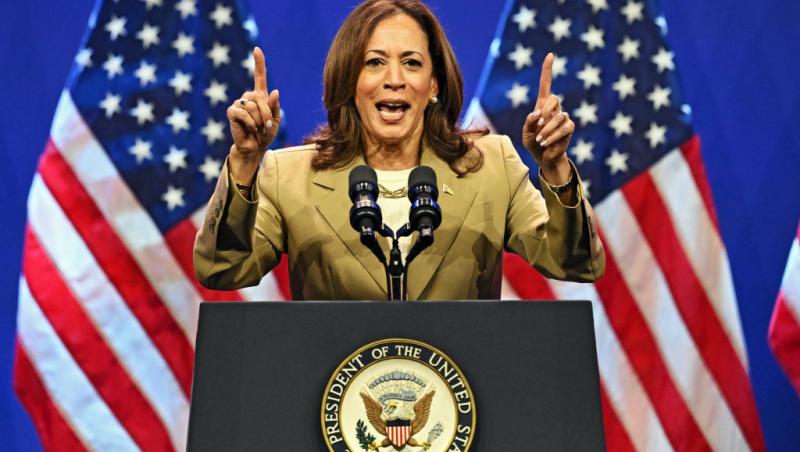 kamala harris la pupitru cu mâinile ridicate, iar în spatele ei două steaguri ale americii