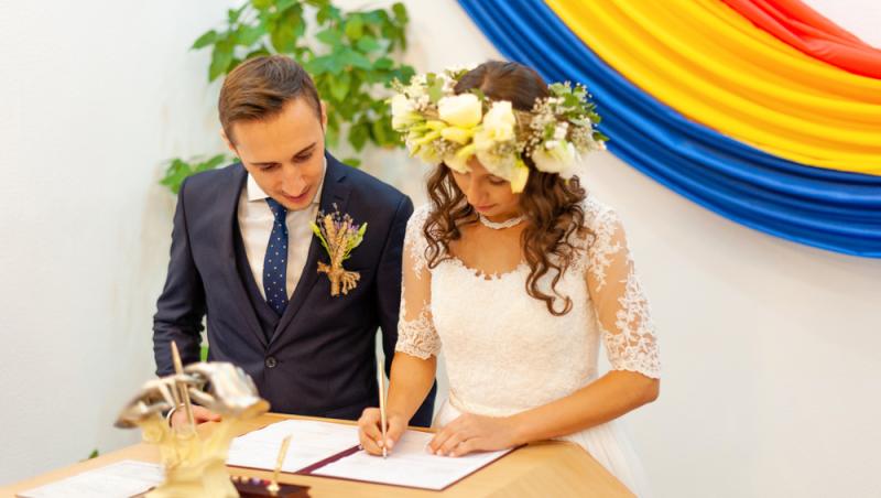 mire și mireasă semnând actul de căsătorie la starea civilă cu steagul româniei în spate