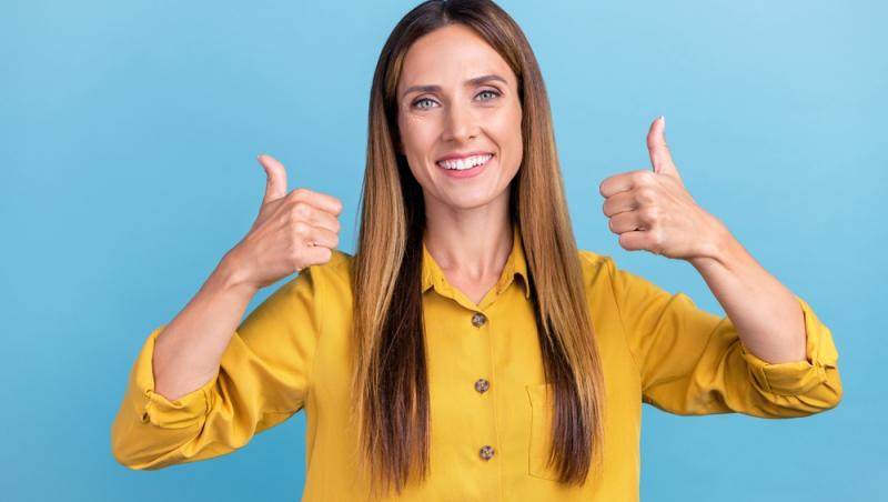 o femeie cu cămașă galbenă care ține degetele mari de la mâini ridicate ca semn de OK, pe fundal albastru