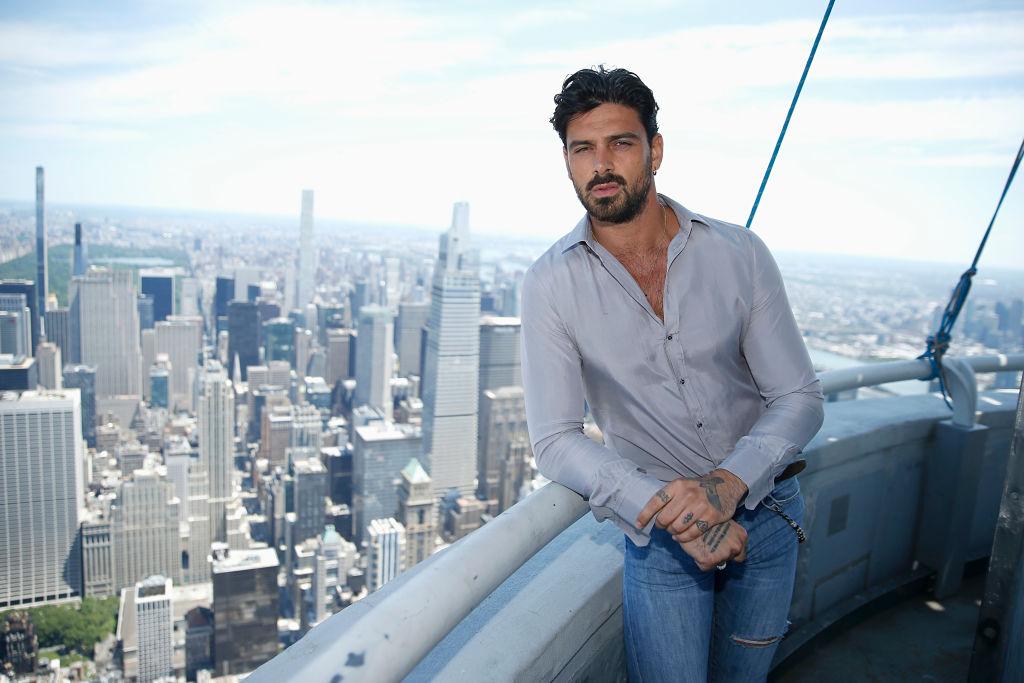 Michele Morrone îmbrăcat casual cu o cămașă gri și blugi rupți, cu tatuajele de pe mâini la vedere