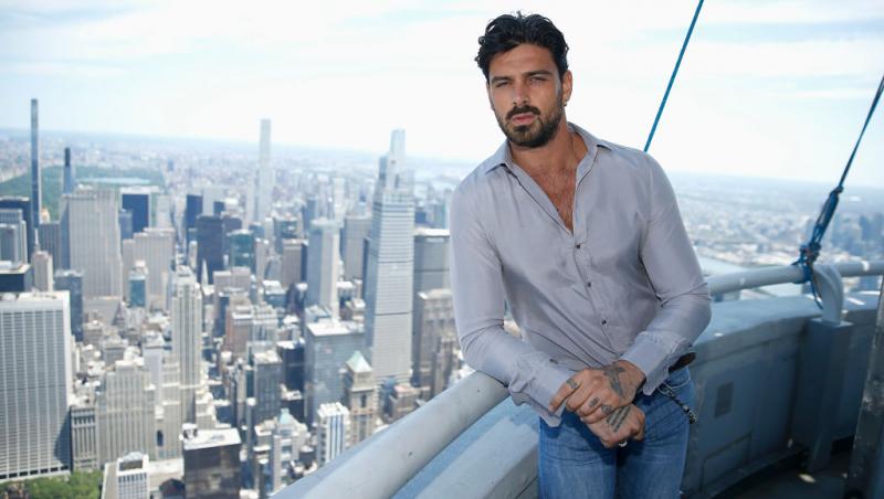 Michele Morrone îmbrăcat casual cu o cămașă gri și blugi rupți, cu tatuajele de pe mâini la vedere