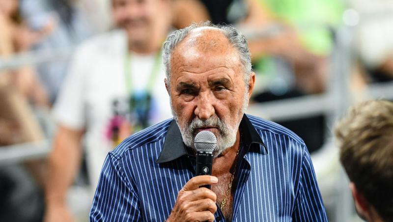 Ion Țiriac îmbrăcat cu o cămașă bleumarin, vorbind la microfon