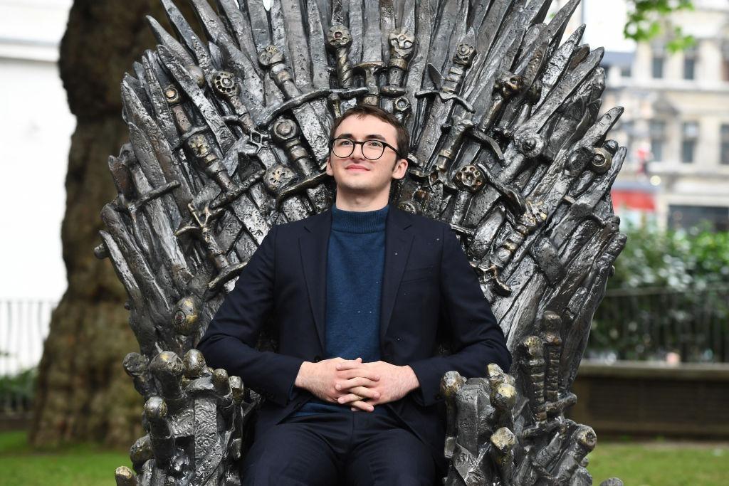 Isaac Hempstead Wright îmbrăcat cu un sacou negru peste o bluză bleumarin, stând pe tronul din serialul Game of Thrones