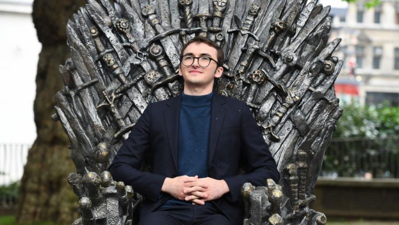 Isaac Hempstead Wright îmbrăcat cu un sacou negru peste o bluză bleumarin, stând pe tronul din serialul Game of Thrones