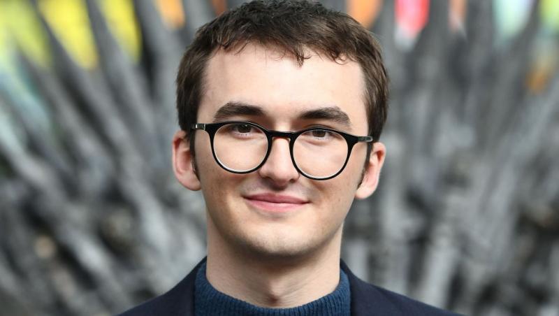 Isaac Hempstead Wright îmbrăcat cu un sacou negru peste o bluză bleumarin, stând pe tronul din serialul Game of Thrones