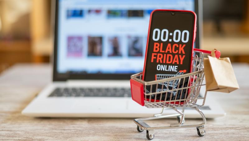 Anunț Black Friday 2025 pe ecranul unui telefon și un laptop pe fundal deschis pe un site de cumpărături