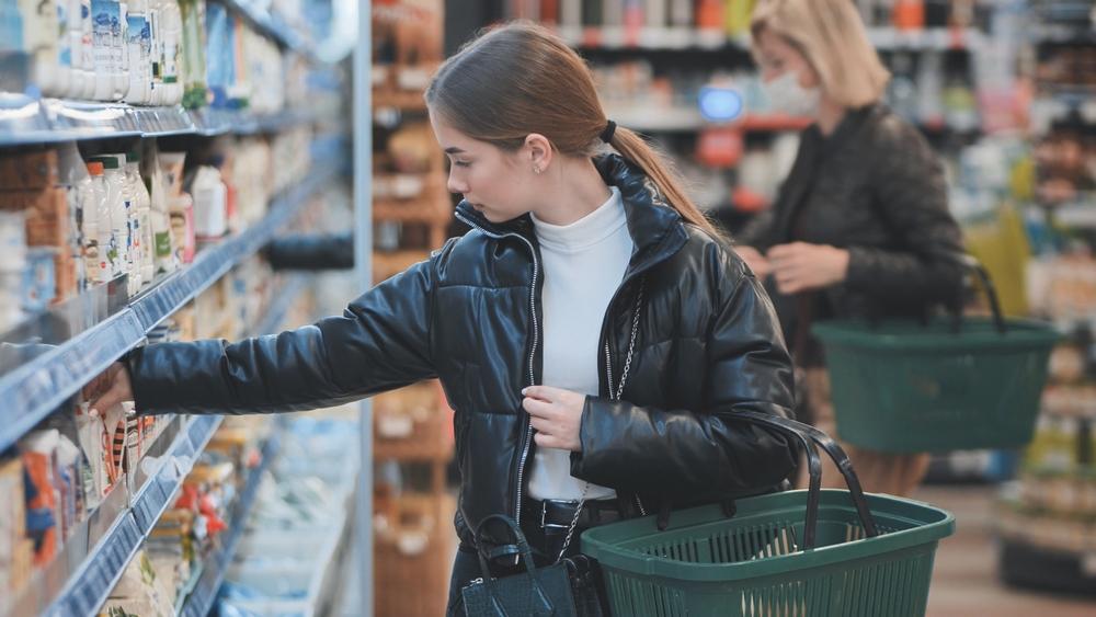 tânără care ține un coș de cumpărături și se uită la produsele de pe raftul unui supermarket