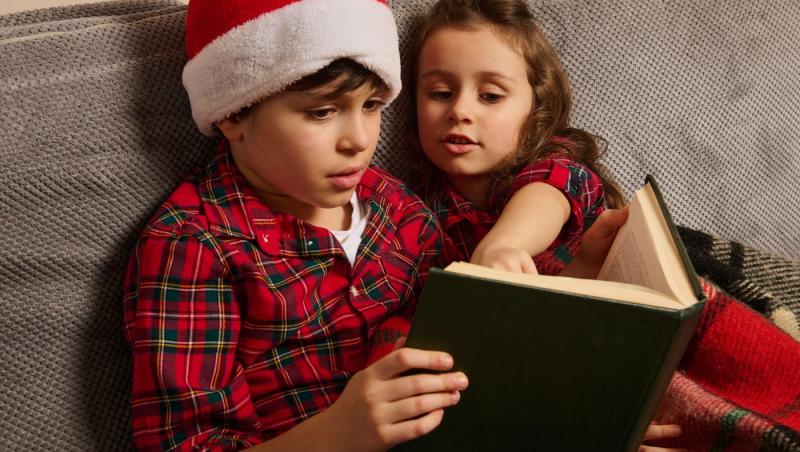 fetiță și băiețel citind dintr-o carte &icirc;n pijamale de crăciun