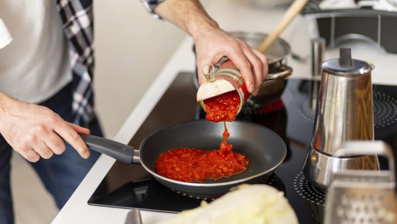 O femeie care toarnă o conservă de roșii &icirc;ntr-o tigaie de pe aragaz