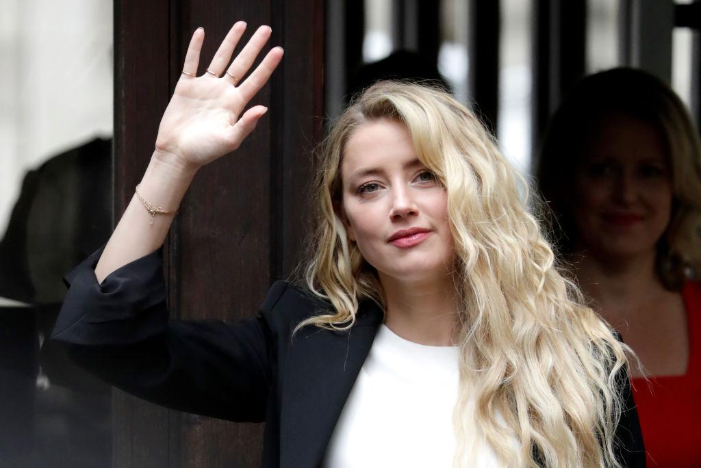 Amber Heard, îmbrăcată într-un tricou alb, peste care poartă un sacou negru, în timp ce salută